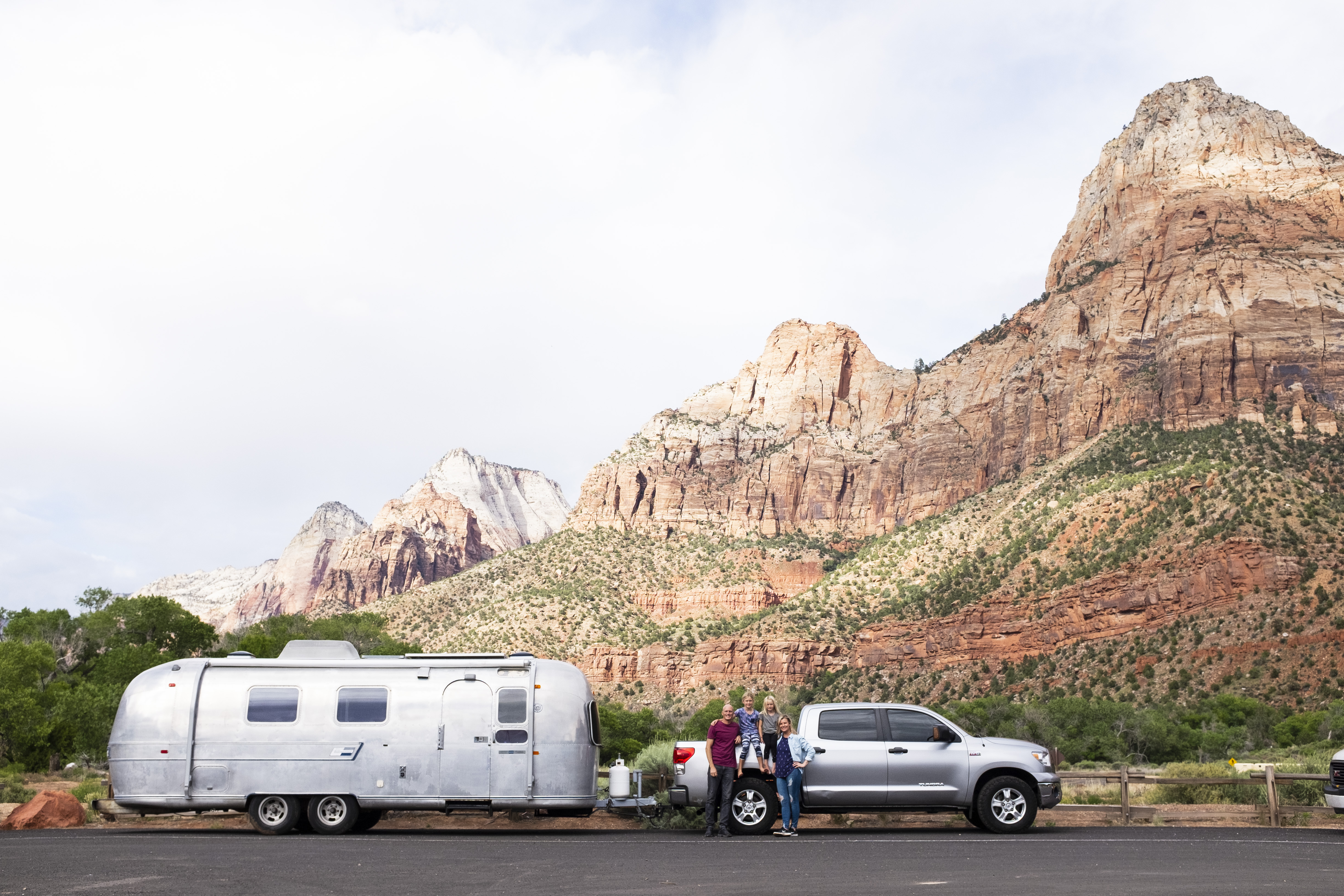Airstream trailer being towed by a silver trunk with a family posing and Zion mountains in the background