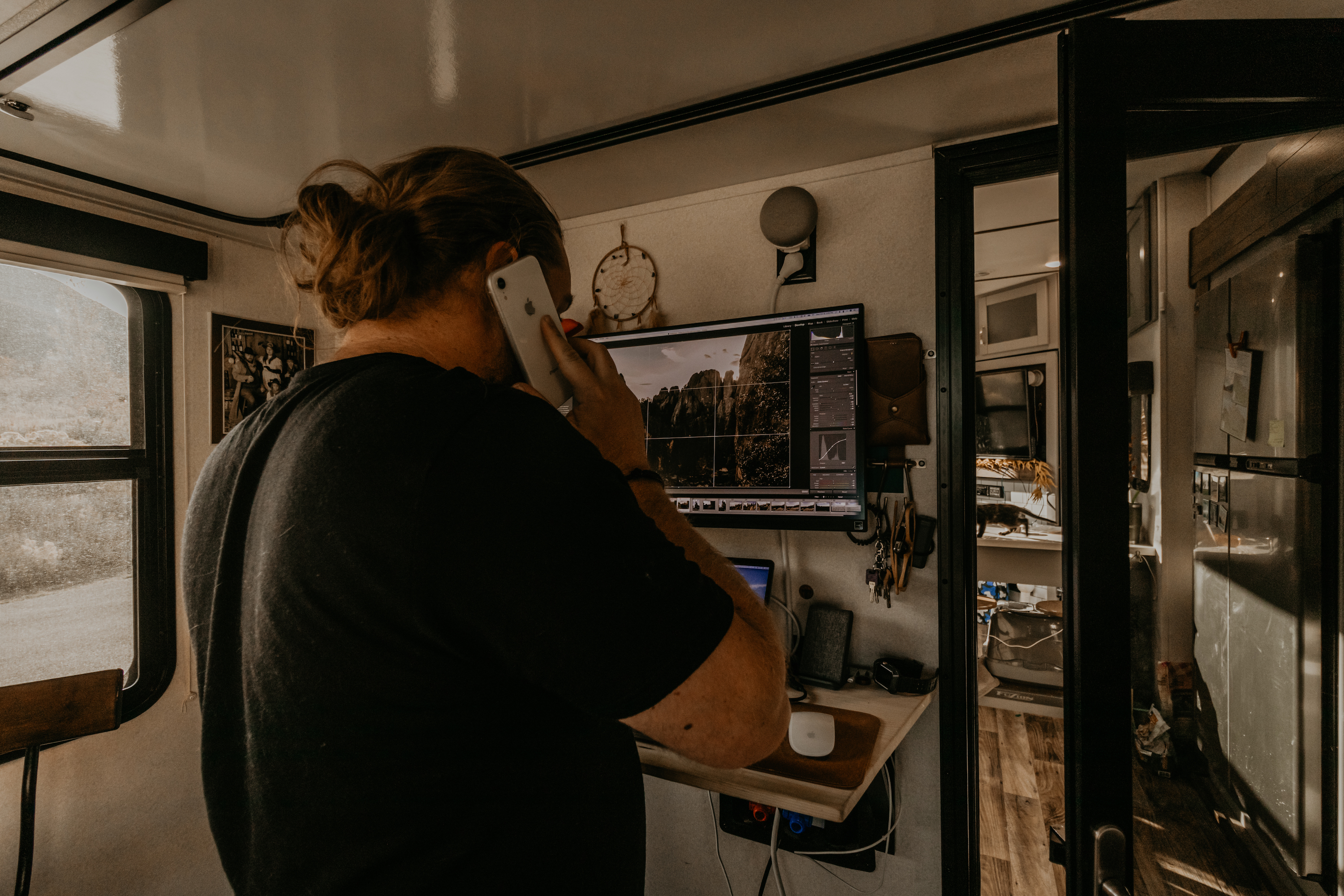 A man standing at a desk, taking a phone call while working on his computer.