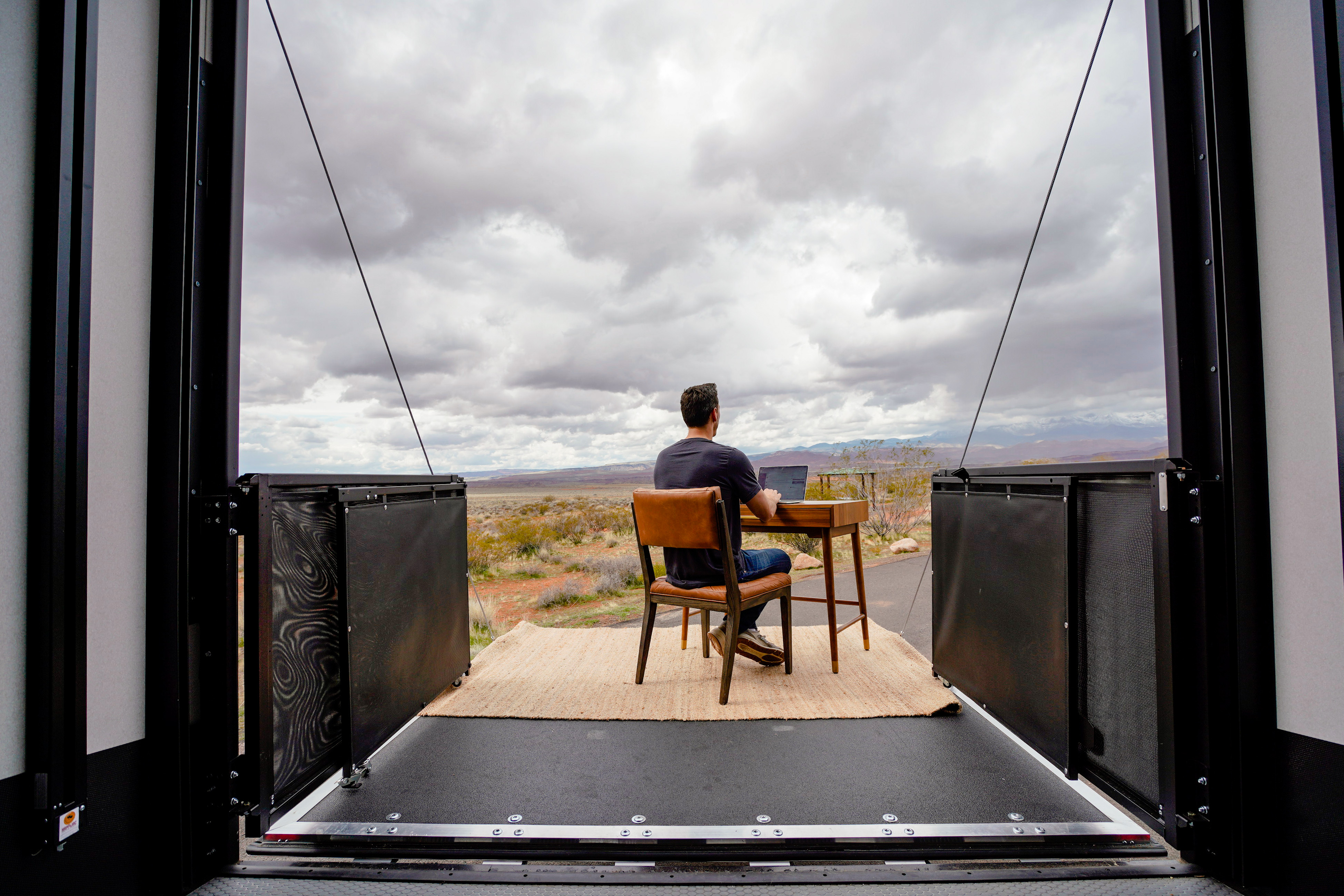 A man sits at a desk on the porch of his toy hauler RV.