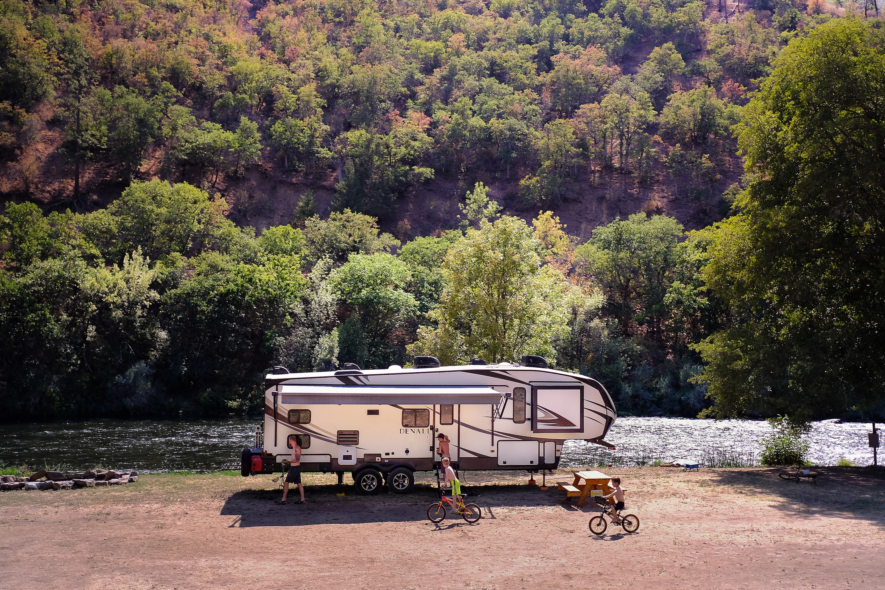 Young boys play and ride bikes in front of an RV near a river.