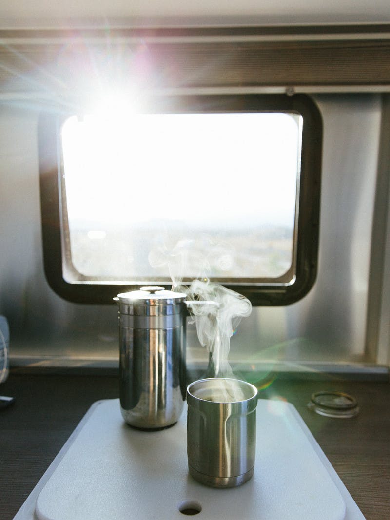Steaming cup of coffee, next to a metal French press, in front of an RV window.