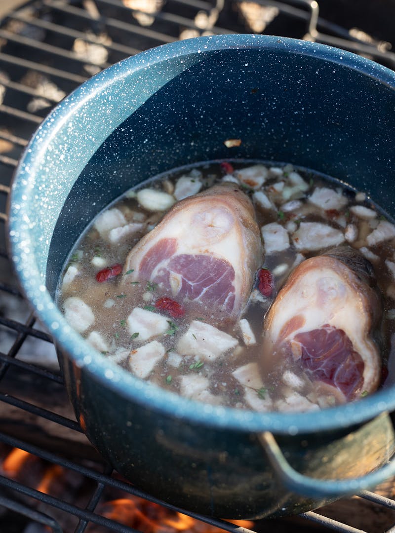An enamel pot over a campfire with ham hocks and beans cooking inside.