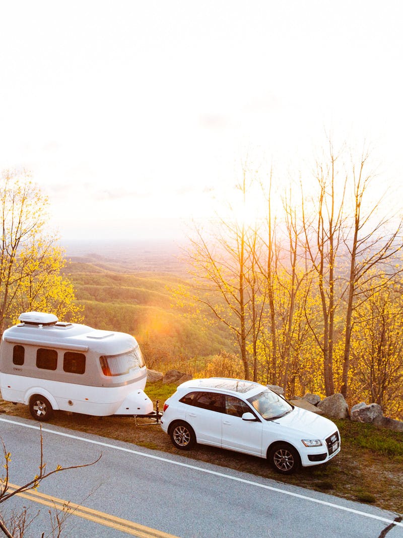 A white car towing an Airstream Nest pulled off on the side of the road with yellow, fall color trees and mountains in the background.