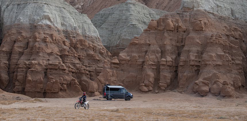 Kalen Thorien ridges a dirt bike in the desert with her TMC Sanctuary Class B RV parked.