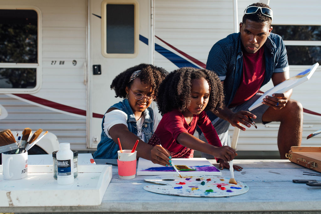 A dad and his two daughters painting at a picnic table outside an RV. 