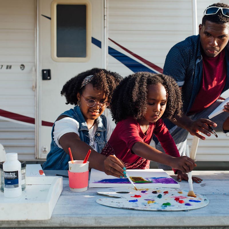 A dad and his two daughters painting at a picnic table outside an RV.