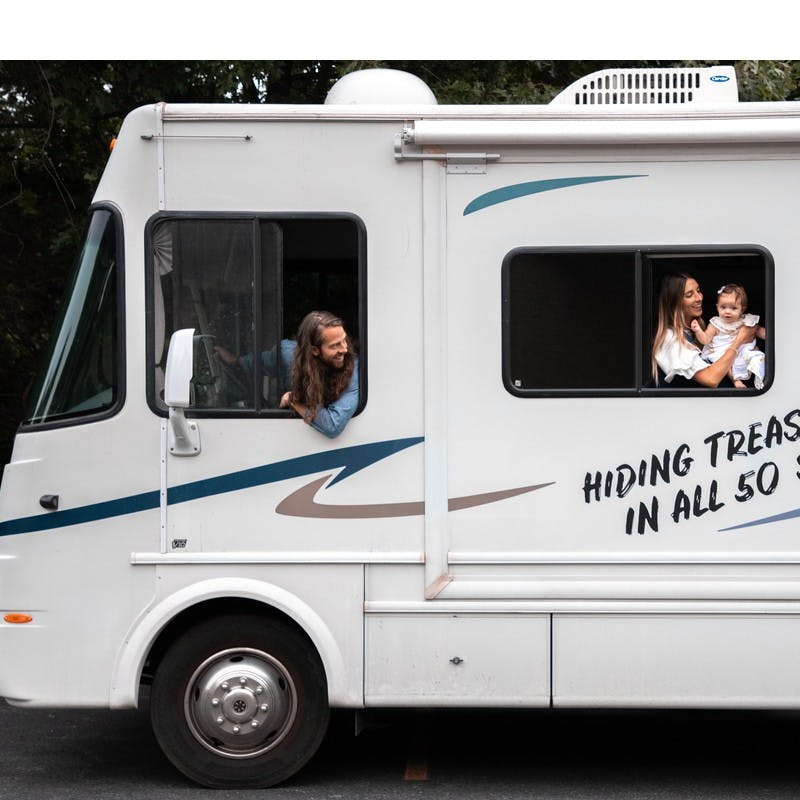 The Jurgy's Class A motorhome, with the man driving, looking out the side window back at the woman holding a baby in the back window.