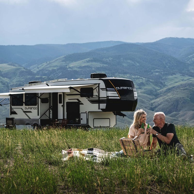A couple having a picnic in a grassy field surrounded by mountains with a KZ RV Durango.