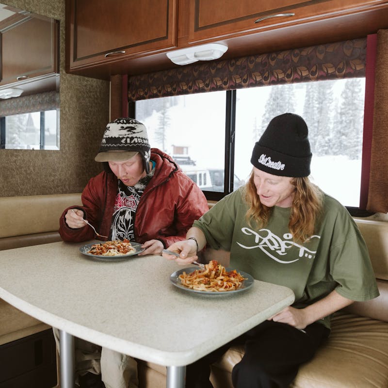 Two young guys in a Class A motorhome sit and eat spaghetti