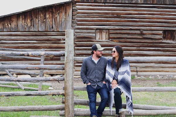 Juli & Jordan Cote stand in front of wooden fence and log barn, Juli with sunglasses and blanket wrapped around her and Jordan with a baseball hat on.