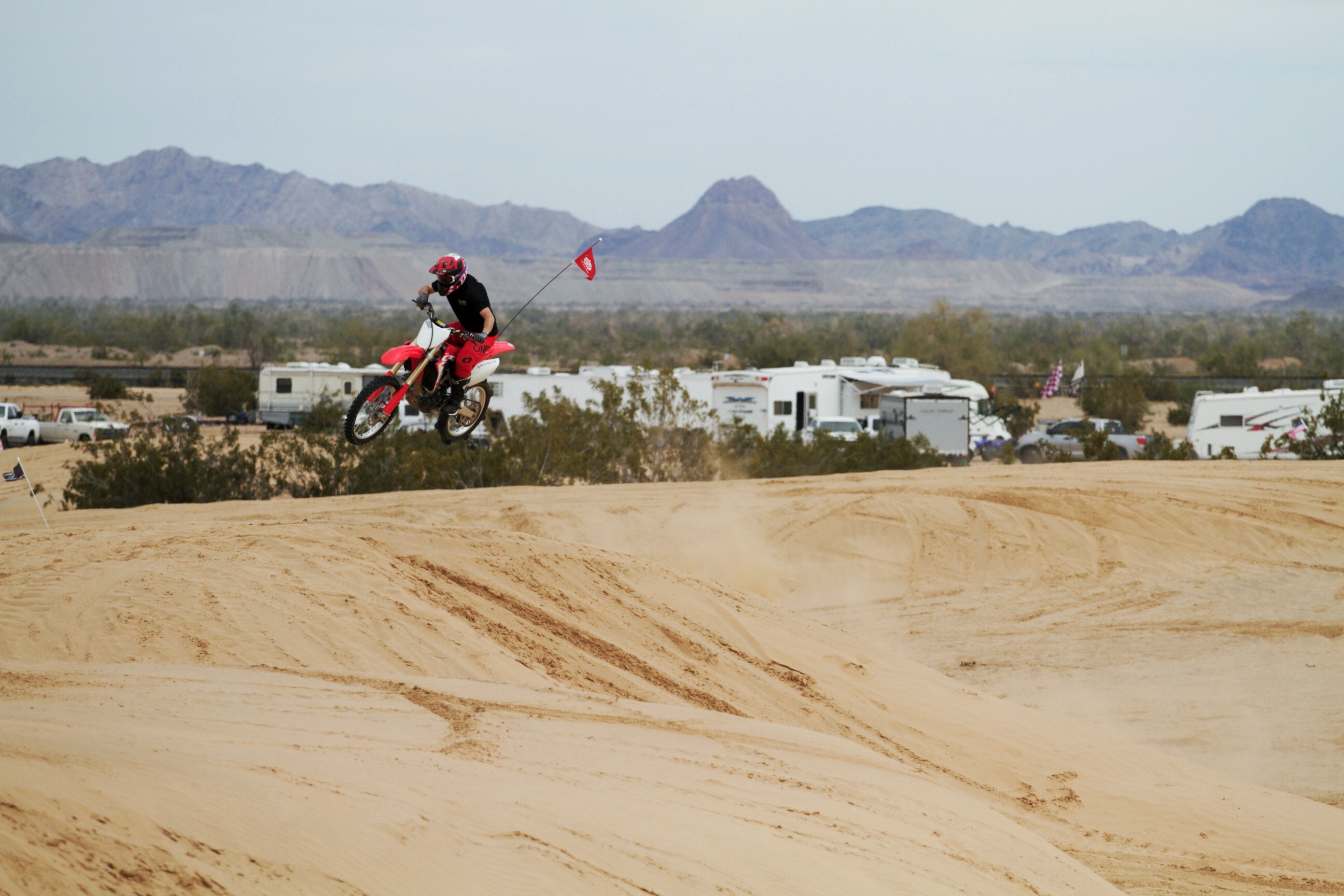 A dirt biker getting air over a sandy dune with RVs in  the distance.