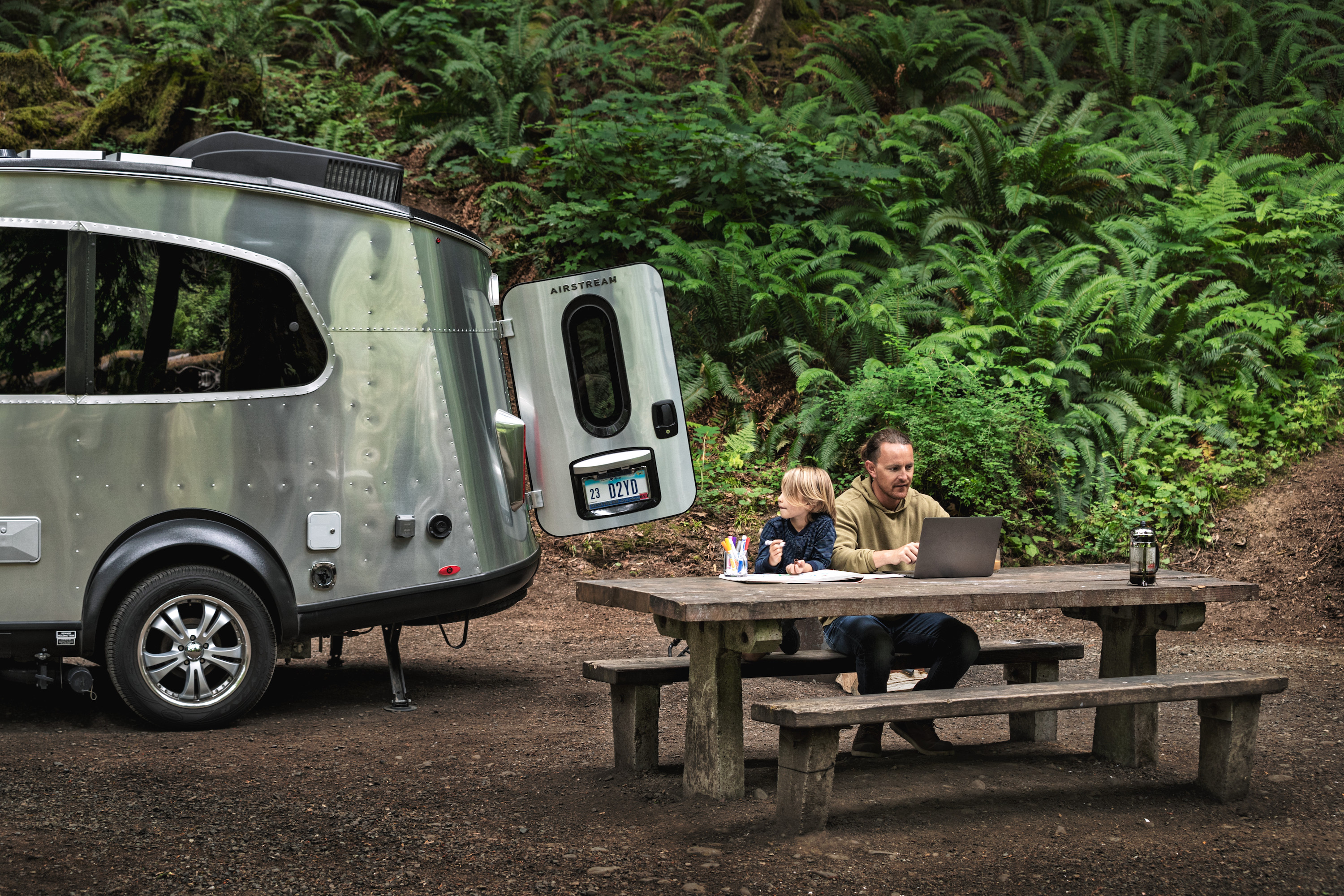 A father works on his laptop at a picnic table while his son colors with an RV next to them.