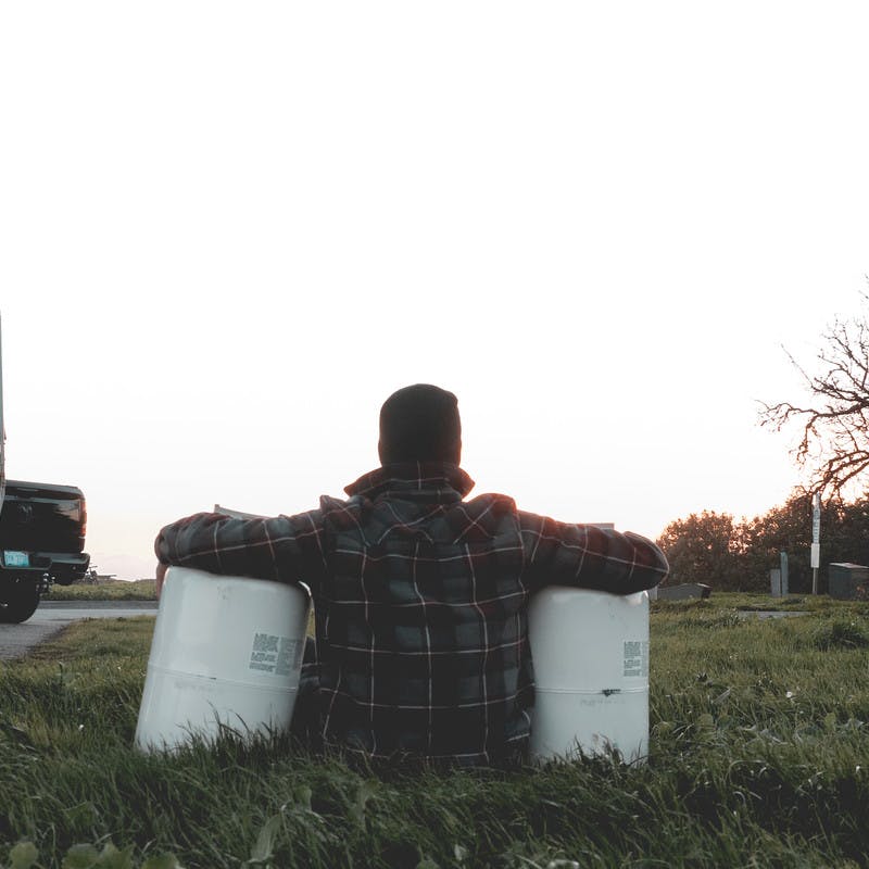 A man sitting in the grass with his arms looped around propane tanks on either side of his body. 