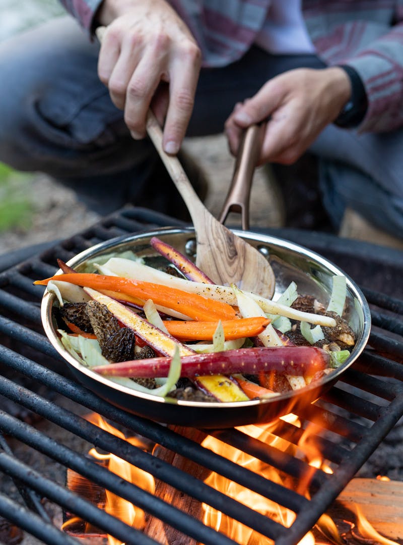 Man in flannel stirs pan with wooden spoon, carrots, morels and fennel cooking over open campfire.