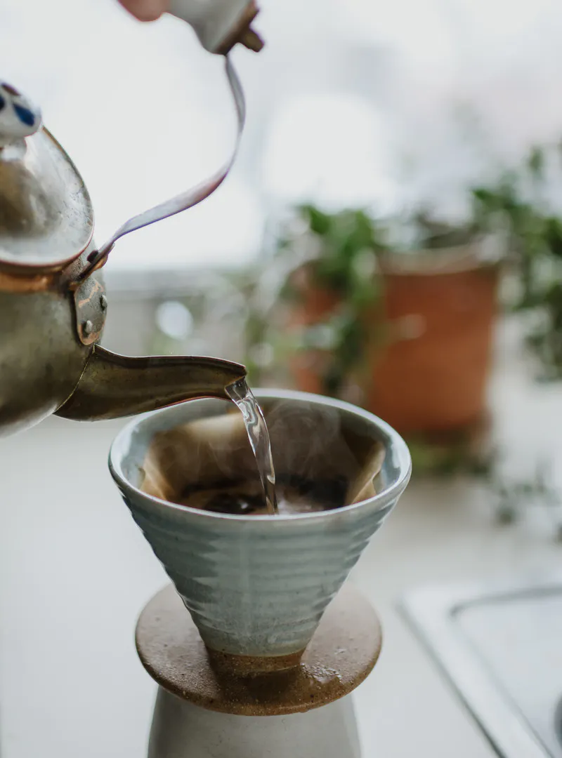 Old copper tea kettle pouring hot water into a coffee pour-over mug, with a plant in the background.