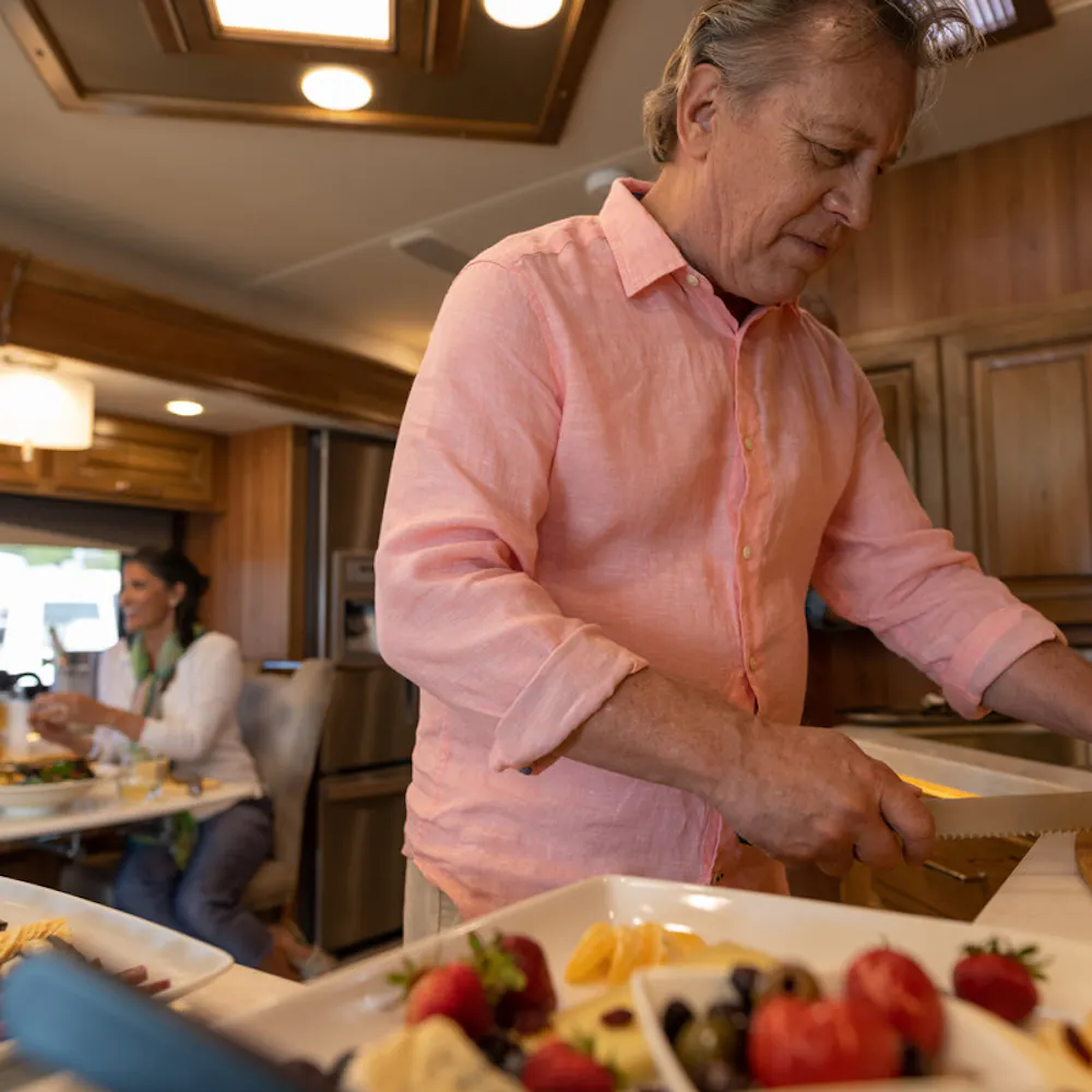 A man slices food in an RV kitchen with fresh fruit and a woman visible in the background.