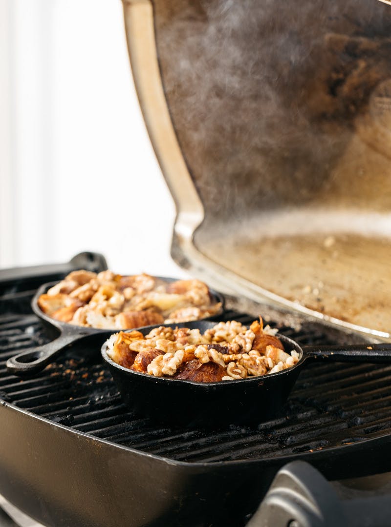 Croissant puddings steaming in two small cast iron skillets placed on a grill grate. 