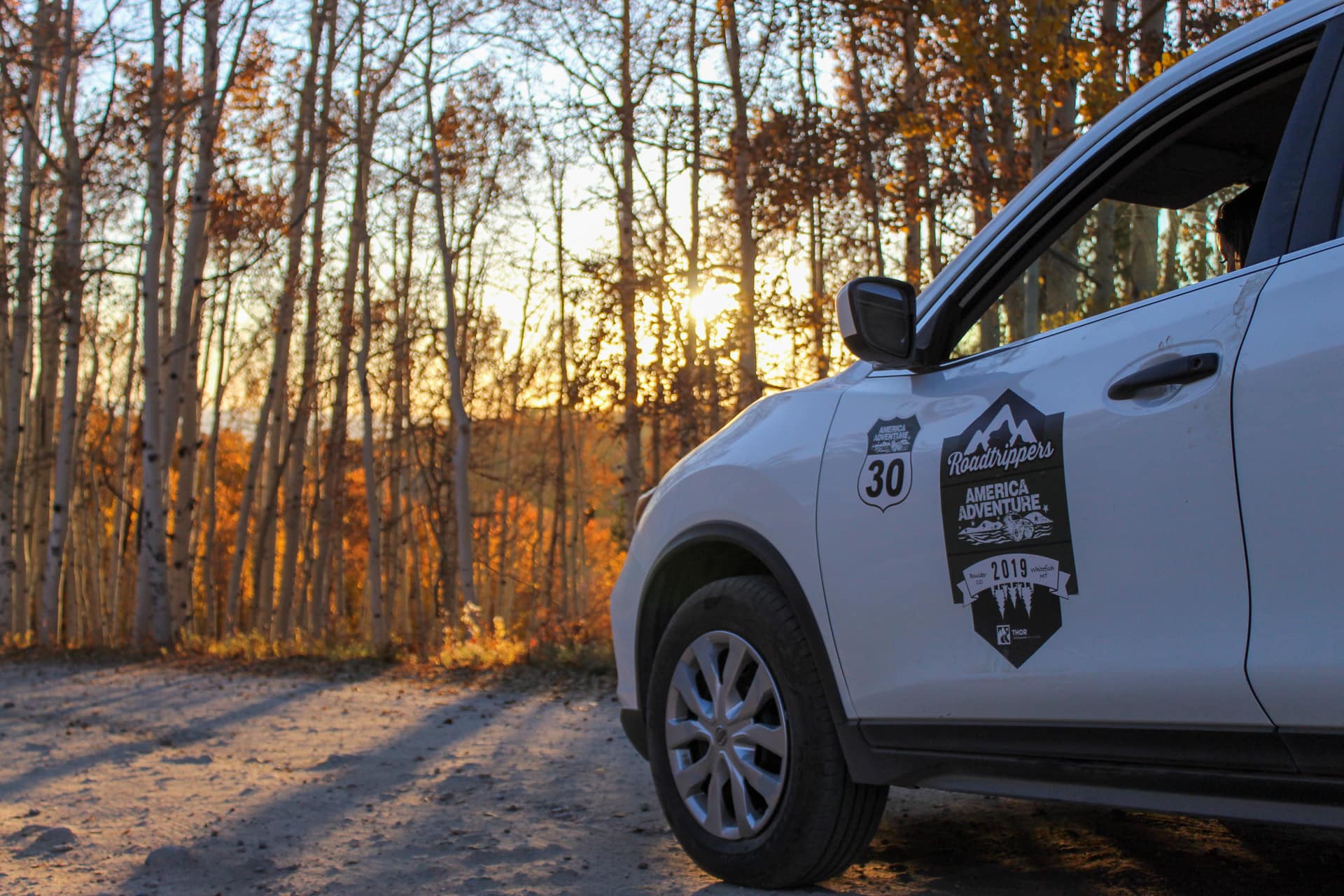 White car with America Adventure decal on dirt road with forest and golden sunset light