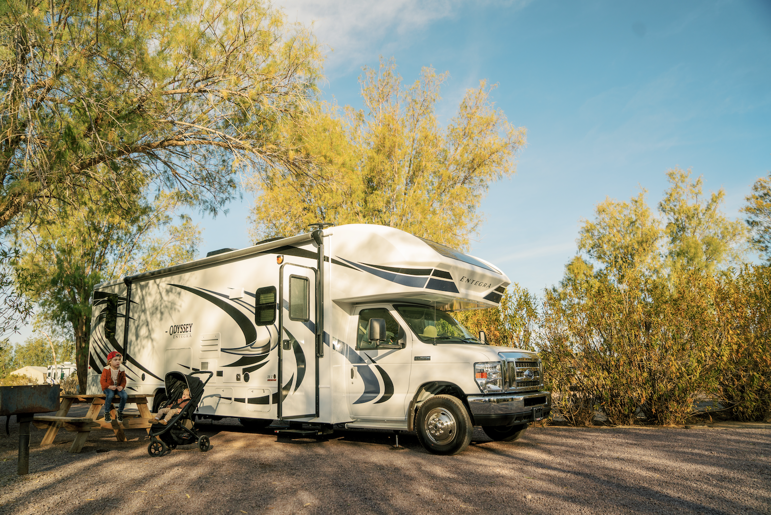 One young boy and child in stroller in front of a Class C motorhome in campground with trees