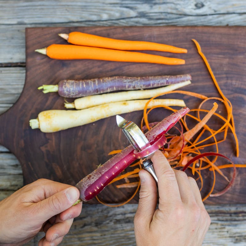 Close up of hands peeling orange, purple and white carrots on cutting board.