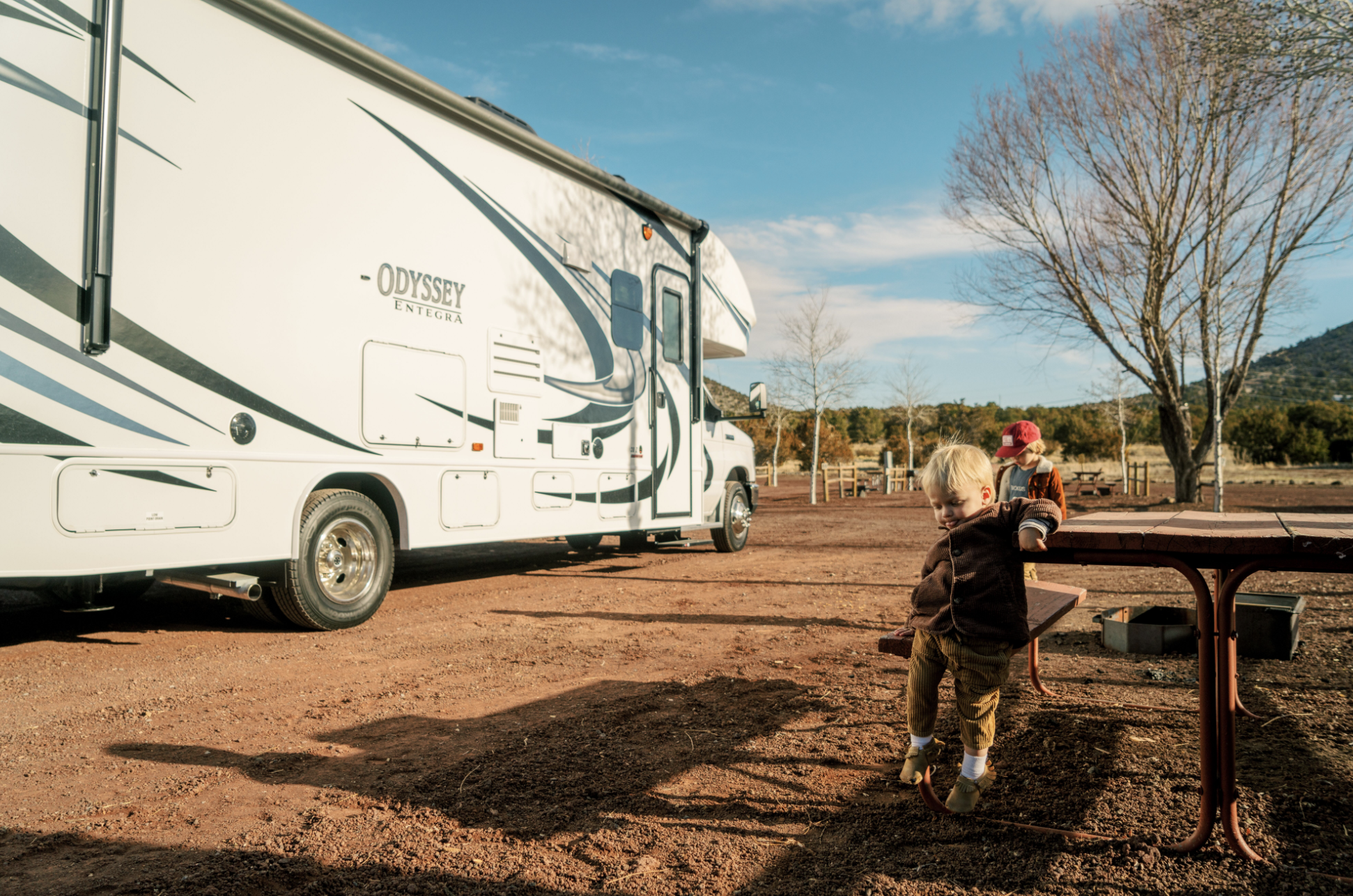Two young boys sit at picnic table at dirt campsite in front of Thor class c motorhome