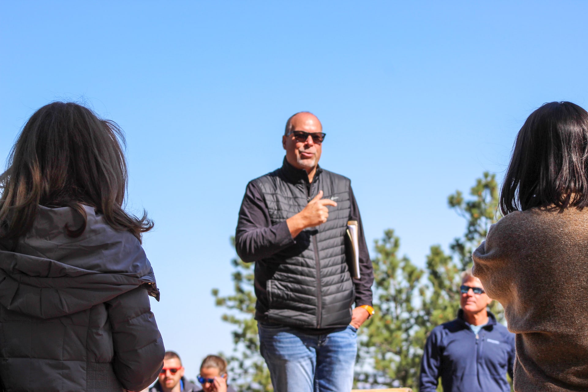 Man in black vest and sunglasses speaks to crowd of people outside