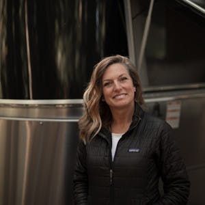 Karen Blue, wearing a black jacket, smiles outside of her Airstream posing for a picture.