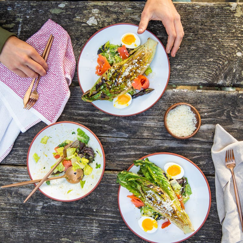 2 caesar salads sit on top of a picnic table while man picks up fork and knife to prepare to eat the salad.