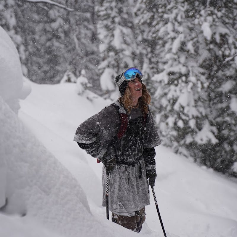 Ryan Barrick with ski poles and ski goggles, surrounded by snow, looking up at the ski and smiling.