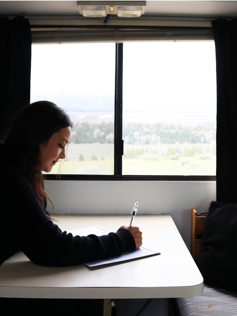 Woman with dark hair sits at the table in her RV and writes in her journal with a view of mountains and trees outside the window.