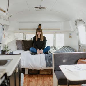 Beth Johnston sitting cross legged inside her RV, on her bed, with gray sheets and a white interior.