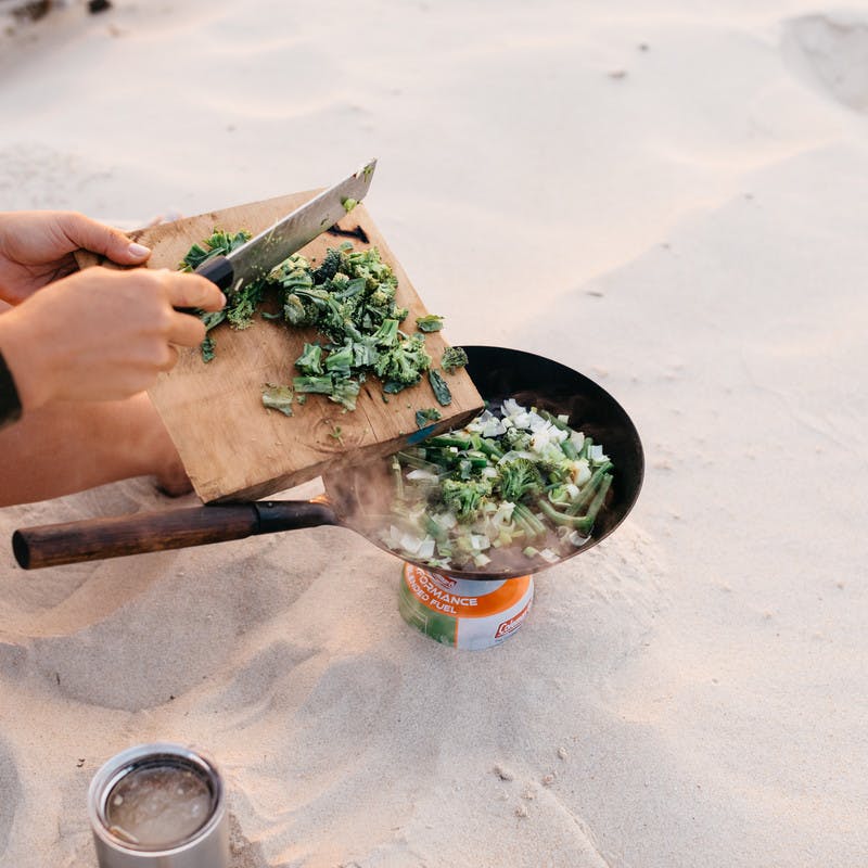 On the beach, a hand scrapes chopped greens off a cutting board with a knife into a cast iron skillet balanced over a small fire.