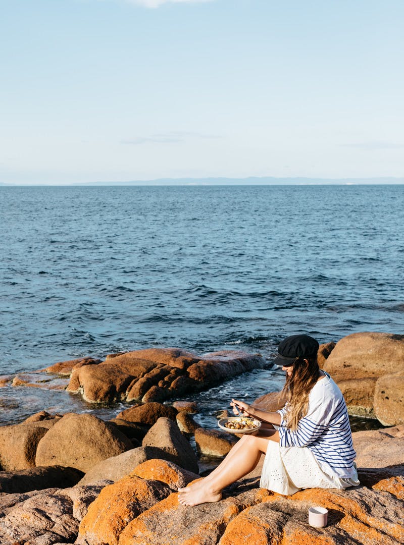 Sarah Glover in a black hat, sitting on orange rocks by the ocean, eating breakfast.