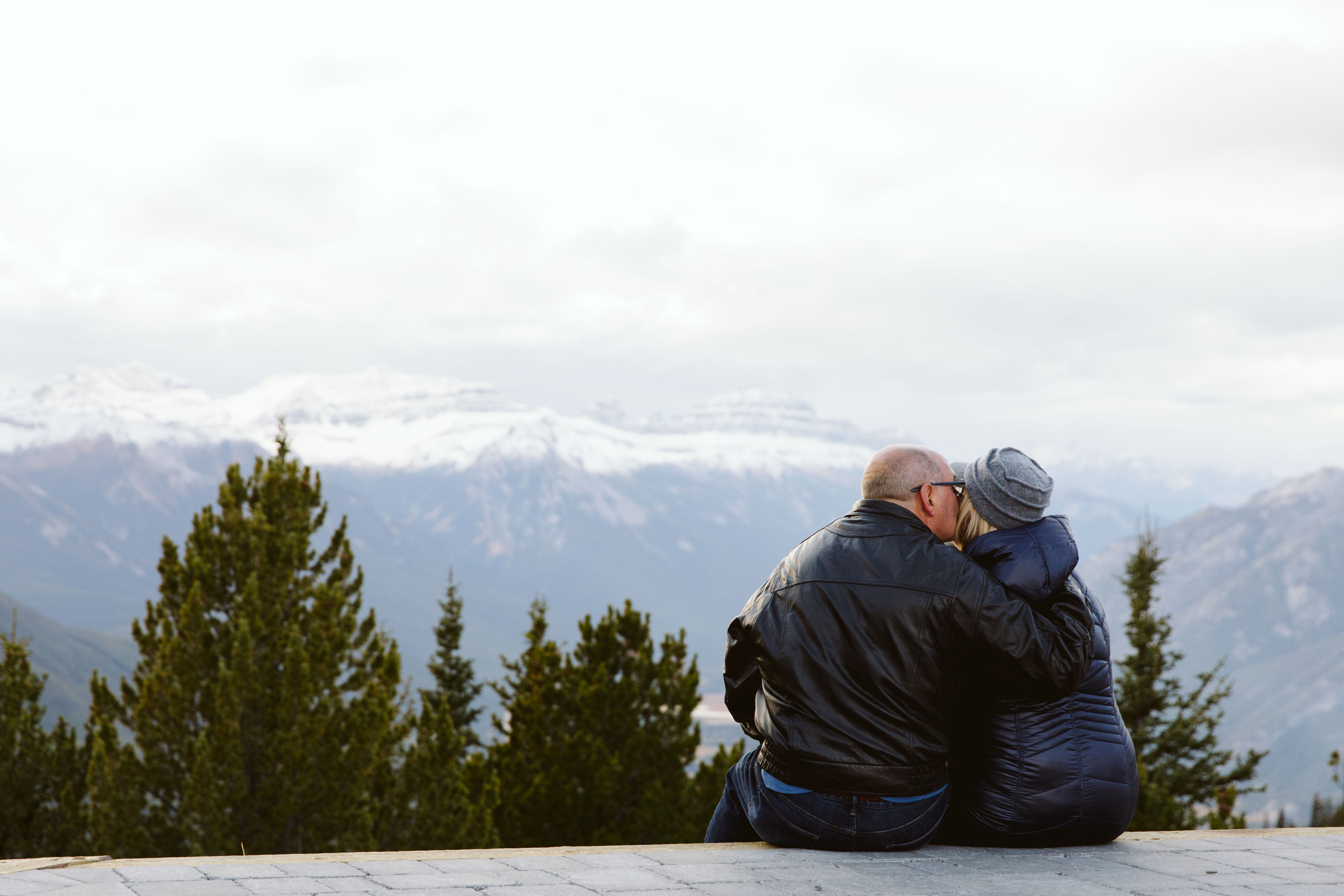 Craig and Tina sitting on a low wall and exchanging a kiss in front of a mountain view. 