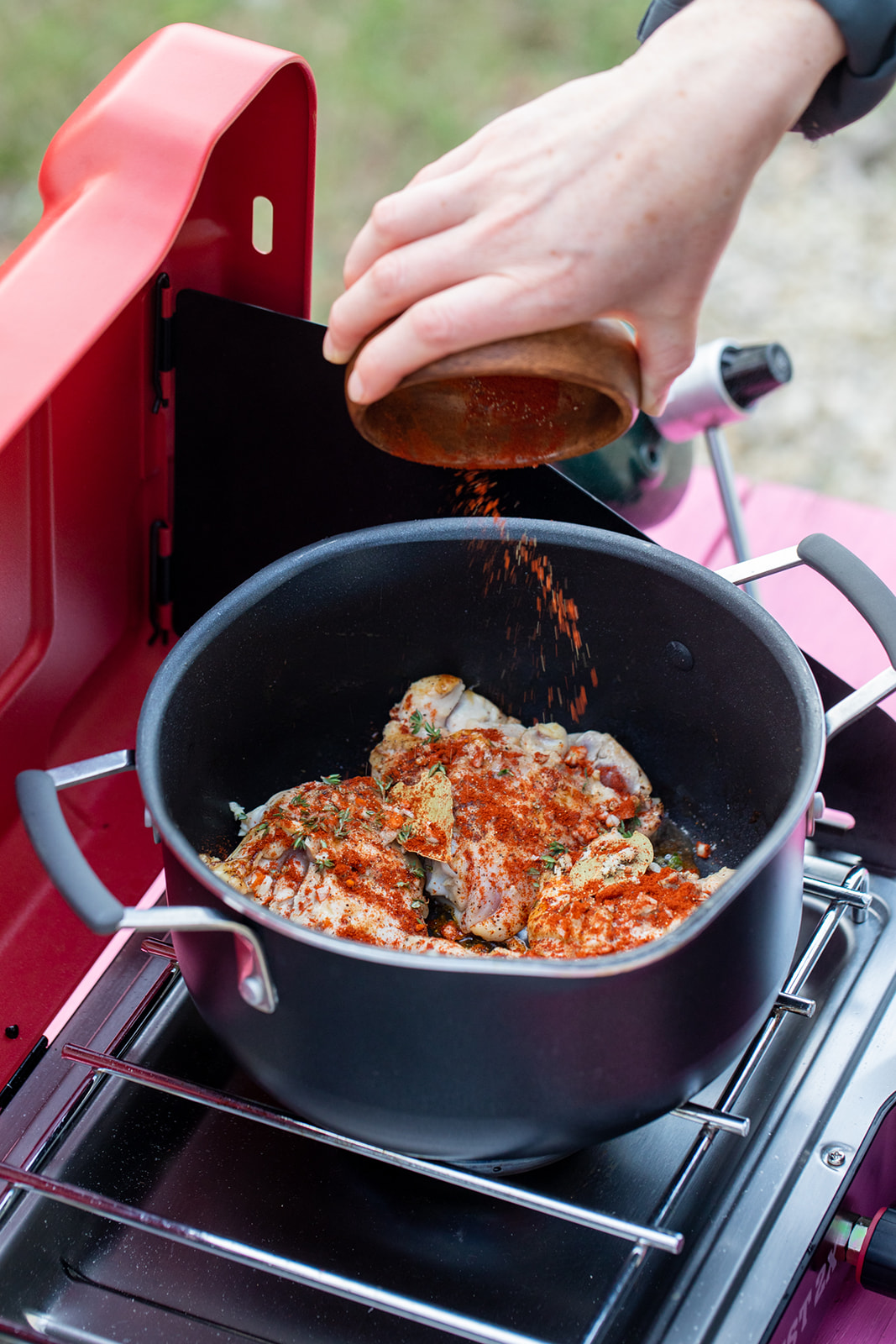 A hand dumping spices into a pot of cooking chicken on a camp stove.