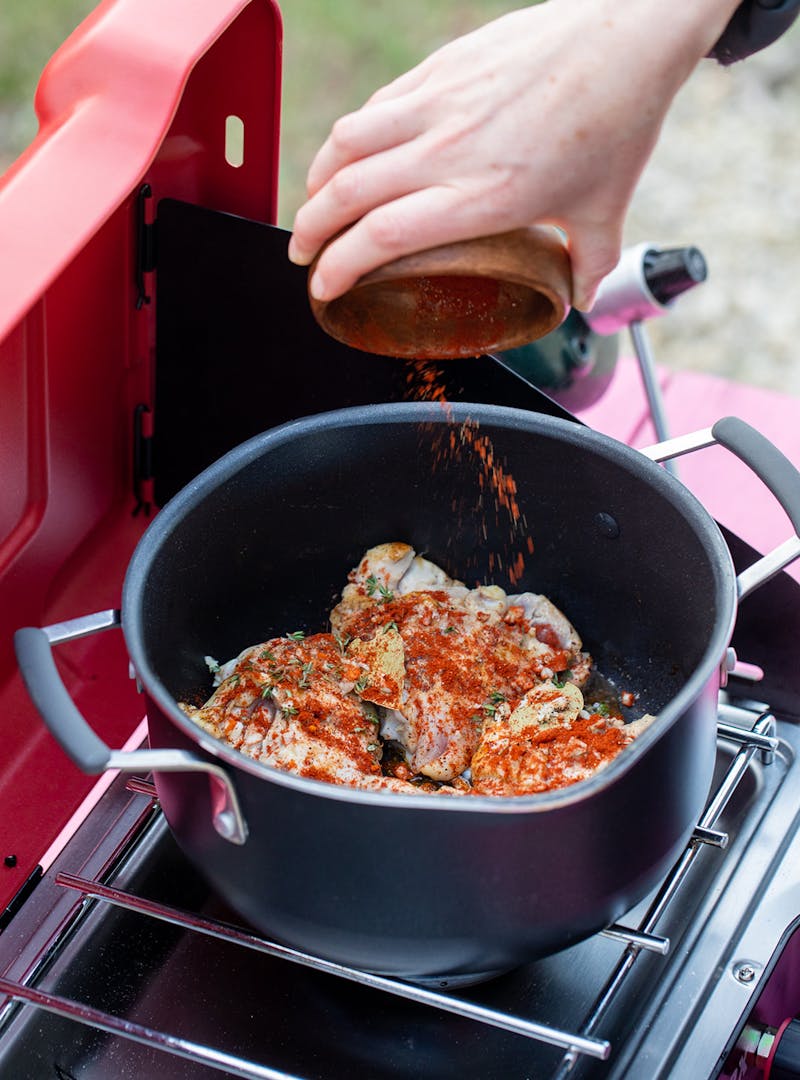 A hand dumping spices into a pot of cooking chicken on a camp stove.