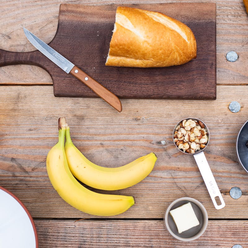 Half cut loaf of bread on cutting board, bananas, hazelnuts, dried cherries, and rum.