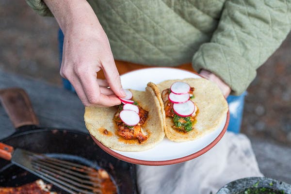 Two blackened rockfish tacos with traditional chimichurri and fire roasted tortillas on a plate, while man tops tacos with radishes. 