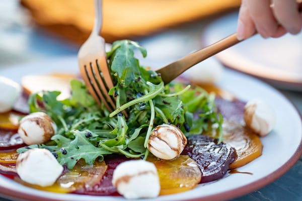 Close up of gold and red beats, topped with arugula, mozzarella cheese balls, and drizzled with a balsamic dressing, includes a knife and fork.