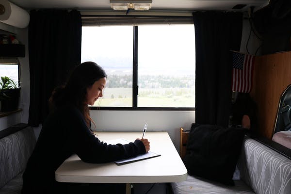Woman with dark hair sits at the table in her RV and writes in her journal with a view of mountains and trees outside the window.