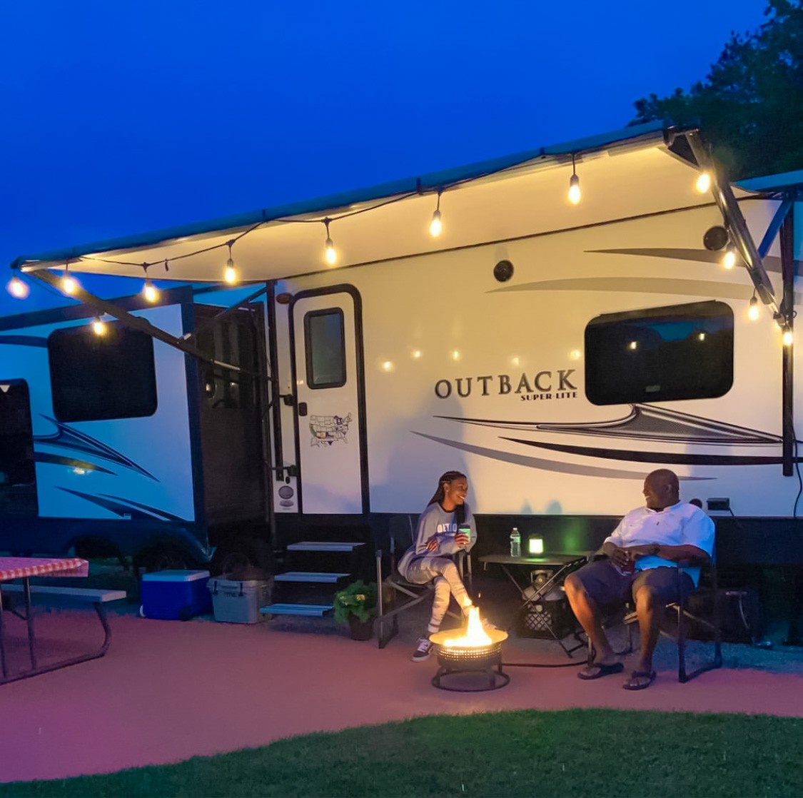 A couple sits in front of their Keystone Outback RV by a campfire at night 
