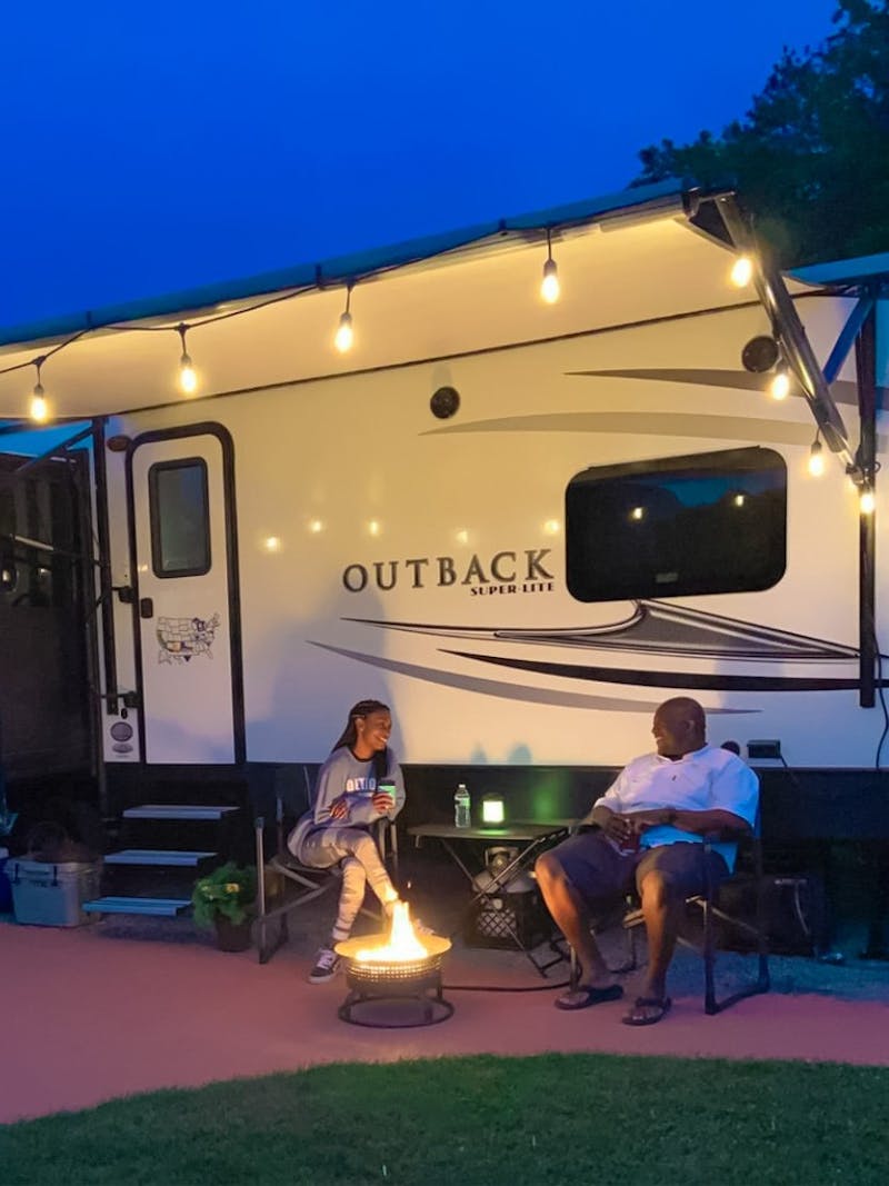 A couple sits in front of their Keystone Outback RV by a campfire at night 