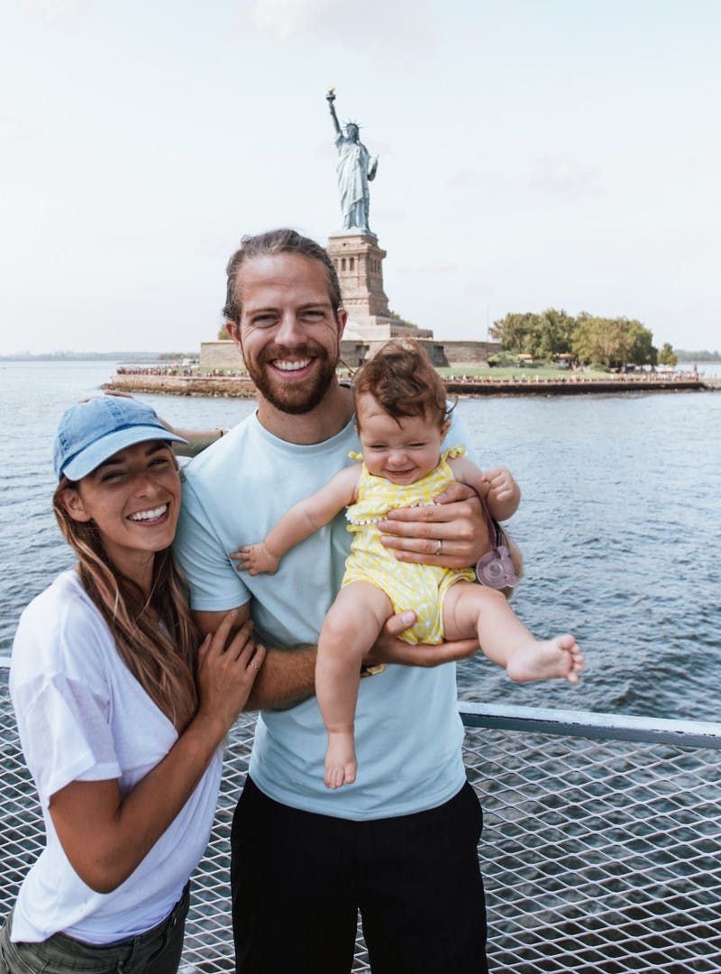 Bryce, Nellie and Avalyn Jurgy, woman and man holding a baby, on a boat, posing in front of the Statue of Liberty.