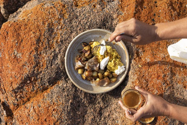 Hand holding a fork over a bowl of eggs, potatoes, and sausage, resting on an orange rock.