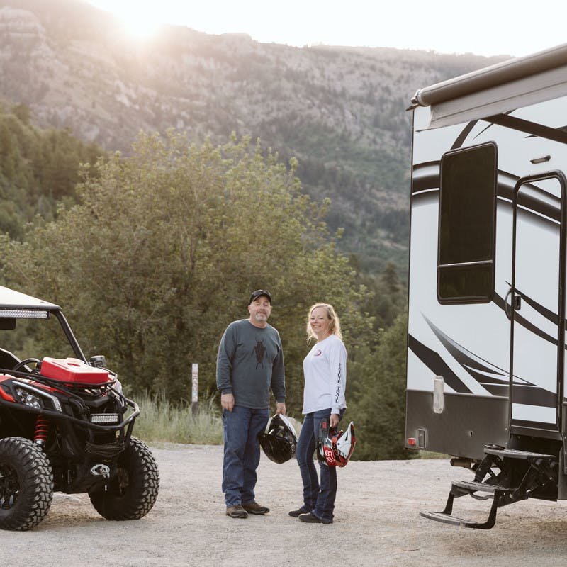  Steve & Suzanne Talbot standing next to an ATV and their Toy Hauler RV with a mountain backdrop. 