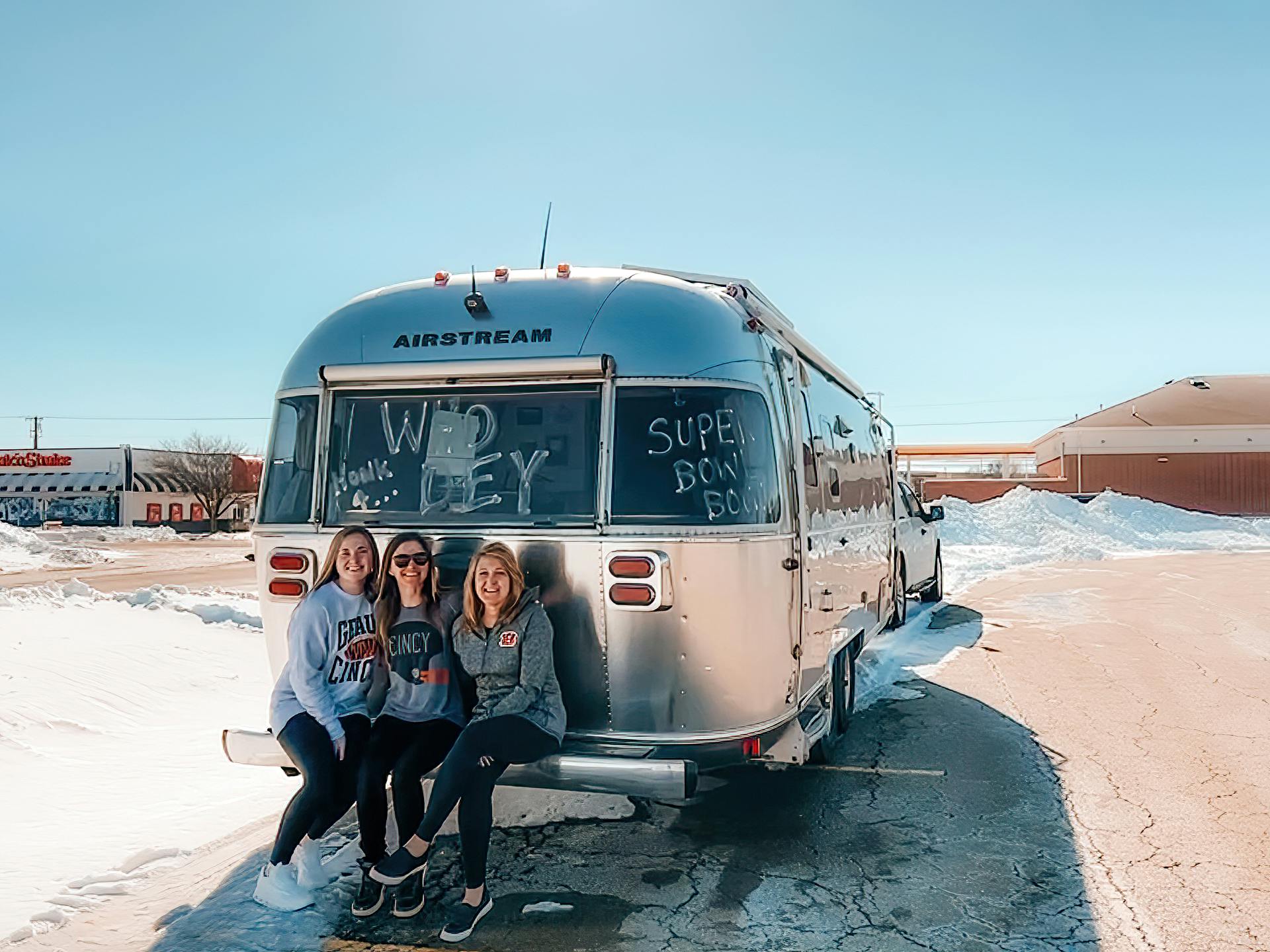 Stacey and her family smiling as they sit on the bumper of her Airstream that is parked near a Steak and Shake 