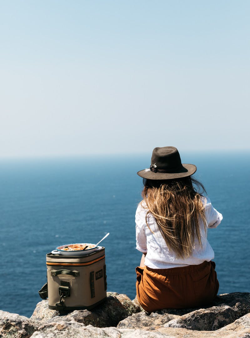 Sarah Glover from the back, sitting on a rock next to a pan of cooked ricotta pasta shells.