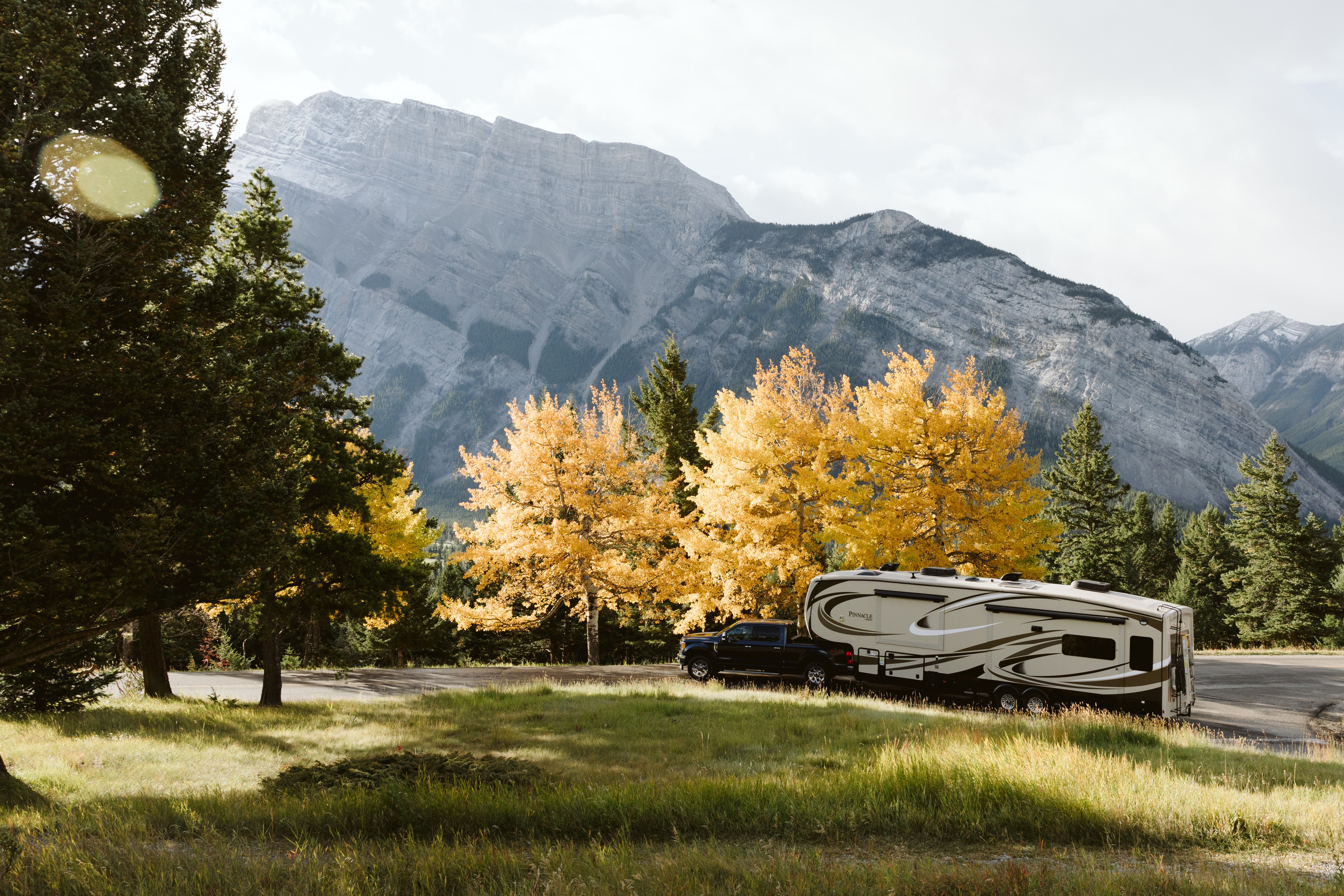 A beautiful nature view with evergreens in the foreground, trees with golden leaves and a mountain in the background, and the truck pulling an RV parked in the middle. 