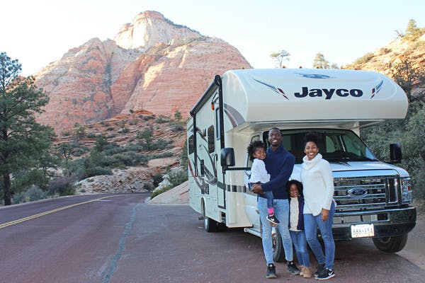An African American family of 4 stands outside of their Jayco RV that is pulled off on the side of the road with the mountains and trees in the background.
