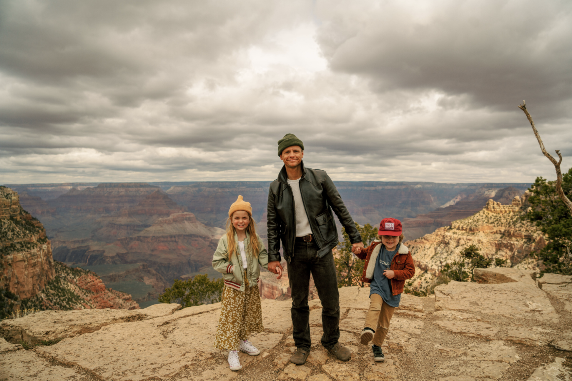 Man in hat holds hands of two young children in front of the Grand Canyon